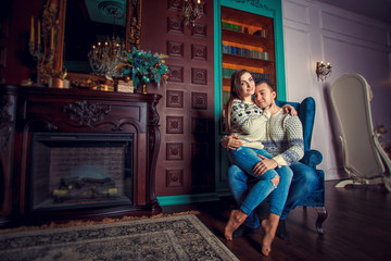 Young couple near fireplace at home. Beautiful stylish young couple celebrates Christmas or New Year near the fireplace in the cozy house. Girl sits on the lap of guy in the chair. Copy space.
