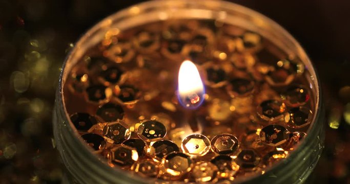 Christmas view. The close shot of a single burning festive candle. Сandle light burns in the dark surrounded by gold sequins. Then the image becomes blurry and turns into spots.