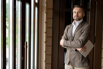 Serious middle aged leader with tablet looking at you while standing by window