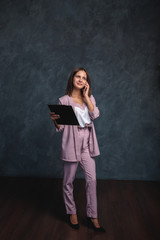 A business girl in a light suit holds a black folder in her hands and talks on the phone. Portrait of a beautiful girl on a dark gray background, space for text. office worker