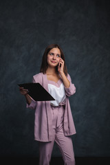 A business girl in a light suit holds a black folder in her hands and talks on the phone. Portrait of a beautiful girl on a dark gray background, space for text. office worker