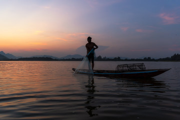 Asian fisherman catching freshwater fish in nature river with fishing Net on wooden boat during twilight. Concept Fisherman's Lifestyle in countryside. Lopburi, Thailand, Asia