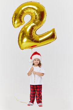 Double Benefit. Portrait Of Little Child Girl In Santa Hat Posing With Gold 2 Two Number Shape Balloon Isolated Over White Background. Christmas, New Year And Birthday Party.