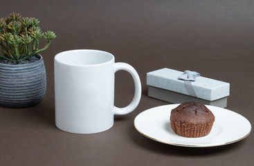 Plain white coffee mug beside cup cake on saucer, flower pot and gift box.