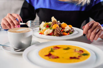 Woman eating business lunch - fresh vegetable salad with grilled meat and pumpkin cream soup in white plates on a white table in a restaurant. Close-up. Space