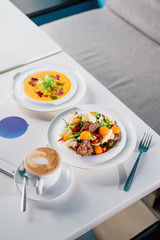 Business lunch - coffee, pumpkin cream soup and fresh vegetable salad with grilled meat in white plates on a white table in a restaurant. Close-up. Space. Flat lay. Top view