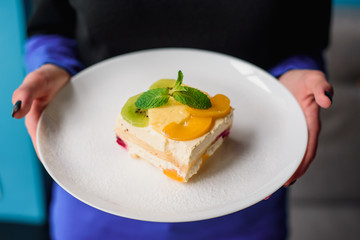 Woman holds a white plate with a slice of fruit cake decorated with a slice of kiwi, pineapple and peach and mint. Close-up. Space.
