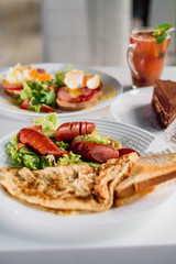 American breakfast - fried sausages, omelet and salad with vegetables on white plate on light table in restaurant. Tea and dessert on blurred background. Close-up. Space. Lunch