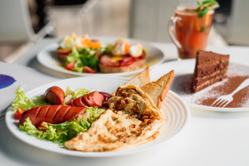 American breakfast - fried sausages, omelet and salad with vegetables on white plate on light table in restaurant. Tea and dessert on blurred background. Close-up. Space. Lunch