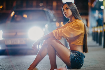 A woman sits on sidewalk against background of night city.