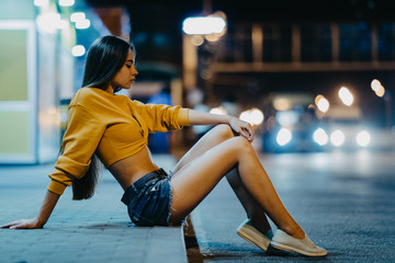 A woman sits on sidewalk against background of night city.