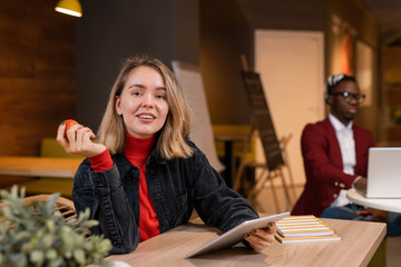 Happy young blonde casual student with tablet and red apple sitting by table