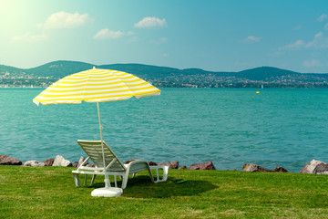 Stylish lounger plastic sunbed with yellow stripes sunshade beach umbrella on the green grass on beach at summer under open sky. Sunbed intended for cold shadow on convenient lounger at Lake Balaton - obrazy, fototapety, plakaty