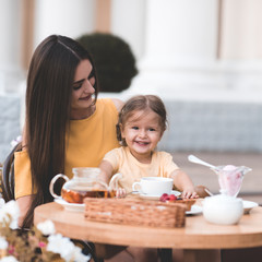 Obraz na płótnie Canvas Happy mother with kid daughter girl 2-3 year old eating cakes in cafe on breakfast outdoors. Good morning. Motherhood. Childhood.