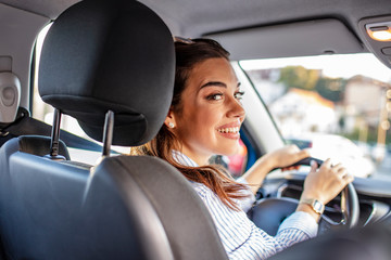 Rear view of attractive young woman in casual wear looking over her shoulder while driving a car. Woman in car indoor keeps wheel turning around smiling looking at passengers in back seat