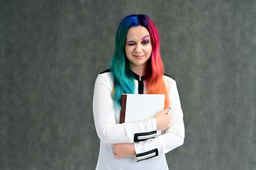 Portrait of a pretty girl in a white shirt with multi-colored hair and make-up on a gray background with a folder in her hands. Stands with a smile in various poses in the studio.