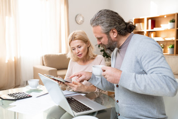 Mature bearded man with cup of tea pointing at document held by his wife