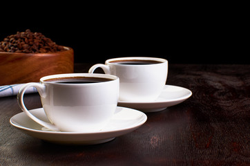 Two cups of coffee in saucers on a dark wooden table. In the background, a wooden dish with coffee beans on a tea towel.