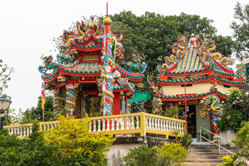 Obraz premium Si Racha, Thailand - March 16, 2019: Red, gold, yellow Wat Koh Loy Buddhist shrines on the hill of Ko Loi Island with gren foliage and under silver sky. Dragon themed decorations.