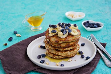 Banana oat pancakes with hazelnuts, sliced banana, honey and honeysuckle on a white plate on a turquoise concrete background. American food. Baking with oatmeal.
