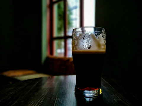 Irish stout in a pint glass on a dark wooden table &ndash; beer is half drunk, light shines through a window