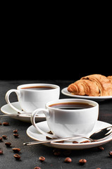 Two cups of coffee on a saucer and coffee beans on a dark background. Croissant in the background.