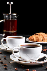 Two cups of coffee on a saucer and coffee beans on a dark background. In the background, a croissant and a coffee pot.