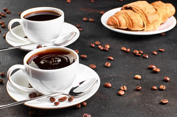 Two cups of coffee on a saucer and coffee beans on a dark background. Croissant in the background. A drop of coffee splashed in the Cup.