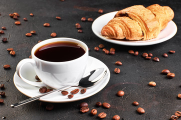 A Cup of coffee on a saucer and coffee beans on a dark background. Croissant in the background.