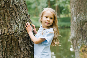 A blonde girl in a blue t-shirt stands near a tree