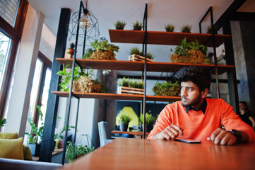 Confident young indian man in smart casual wear like orange sweater, sitting at cafe and using his mobile phone.