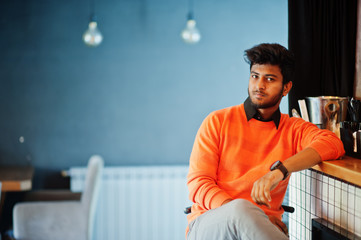 Confident young indian man in orange sweater sitting bar counter at cafe.