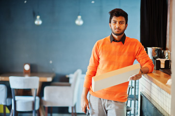 Confident young indian man in orange sweater standing near bar counter at cafe and hold box for delivery with pizza.