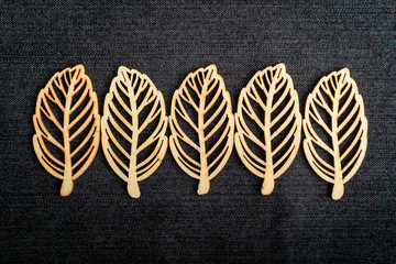 Line with five delicate light brown wooden leaves on dark grey textile material background, top view with space for text on the right side, flat lay with laser cut wooden objects