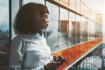A dazzling young African-American woman entrepreneur with a cellphone in her hands is standing next to a panoramic window of a modern office skyscraper with orange lines and pensively looking outside - Powered by Adobe