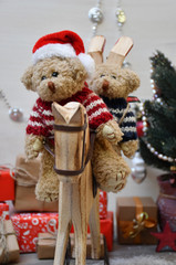Two Teddy Bears are riding a wooden hobbyhorse on a background of a Christmas tree & boxes with Christmas presents
