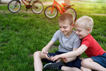 Naklejka na ściany i meble Two boys are gaming on the smartphone while sitting on the grass. Bicycles in the background
