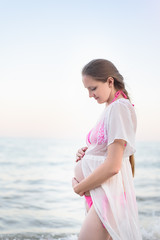 Young pregnant woman is standing on the seashore and hugging her belly. Enjoying the moment.
