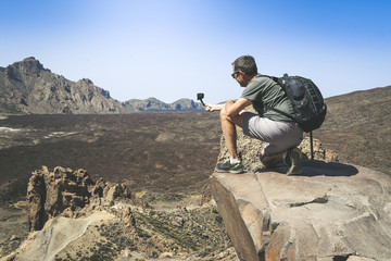 Travel blogger with backpack making video with action cam for social blog. Caucasian man taking pictures on a mountain cliff overlooking the lavic panorama. Holiday, travel, new trend tech concept.