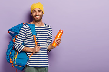 Satisfied unshaven man holds thermos and big rucksack, ready for adventurous trip, smiles...