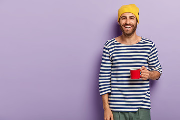 Smiling young man holds red mug, drinks hot beverage, wears yellow hat and casual striped sweater,...