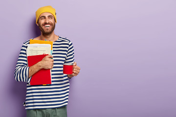 Delighted unshaven young man student holds papers and red notepad, holds cup with hot drink, has...