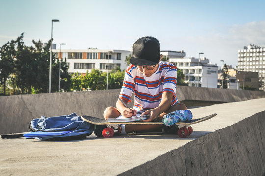 Trendy Student Doing Homework Out Of The College Sitting On A Concrete Wall. Young Boy Writing On An Exercise Book Using A Skateboard Like A Desk In A Urban Context. Educational School,  Youth Concept