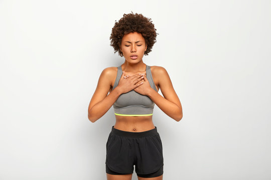 Horizontal shot of dissatisfied Afro American woman suffers from asthmatic fit, breathers deeply, has shortness of breath or dyspnoea, wears grey top and shorts, isolated over white background