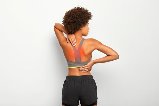 Spine pain or backache concept. Horizontal shot of slim dark skinned woman has cronic painful feelings in back, poses against white background, has injury or cramps, wears grey top and shorts