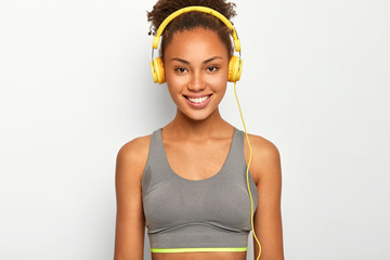 Indoor shot of self determined curly Afro American sportswoman chooses playlist for cardio workout, smiles pleasantly, wears casual top, looks directly at camera, poses over white background.