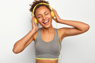 Fototapeta na wymiar Emotional beautiful Afro American woman with curly hair, laughs positively, enjoys loud music in headphones, keeps eyes closed from pleasure, wears grey top, goes in for sport with favourite track