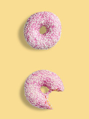 Two pink donuts on a yellow background. One donut is whole and the other is bitten. Donuts with pink glaze and white sprinkles.