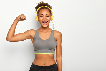 Horizontal view of dark skinned woman in good mood, raises arm with muscles, has strong body, dressed in gym outfit, listens audio via modern headphones, poses indoor. Fitness and music concept - obrazy, fototapety, plakaty
