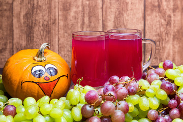 Two glasses of red stum with some green and red grapes and a funny pumpkin photographed in front of a wooden wall.